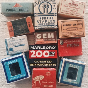 Puede incluir: Una colección de cajas vintage que contienen diversos artículos de oficina, como grapas, chinchetas, sujetapapeles y cuchillas para rascar. Las cajas están hechas de cartón y tienen diseños y textos coloridos.