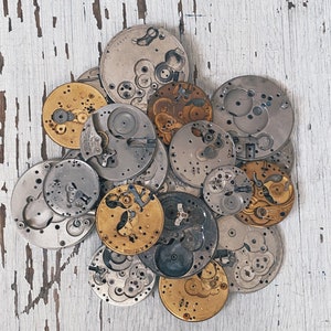 Puede incluir: Una colección de piezas de relojes vintage, incluyendo engranajes, muelles y otros componentes, dispuestos sobre una superficie de madera blanca. Las piezas son de metal y tienen un aspecto desgastado y antiguo.