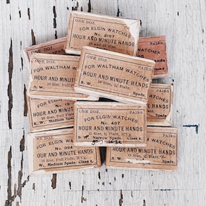 May include: A stack of vintage boxes for watch parts, labeled "One Doz. For Elgin Watches Hour and Minute Hands" with different sizes and styles. The boxes are made of brown cardboard and have white lettering.