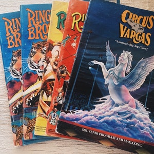 Peut inclure: Quatre livrets de programme de cirque vintage. Le livret du haut est bleu avec le texte "Circus Vargas" et "America's Big Top Giant". Les trois autres livrets sont de Ringling Bros. et Barnum & Bailey Circus. Les livrets sont sur une surface en bois blanc.