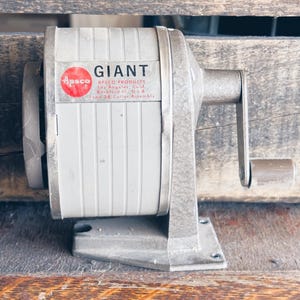 May include: A vintage gray pencil sharpener with a hand crank. The sharpener has the word "GIANT" printed on it, along with the "Apsco" logo and product information. The sharpener is made of metal and sits on a wooden surface.