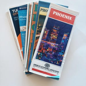 May include: A stack of folded paper maps with a colorful illustration of a city skyline on the top map. The top map is titled "PHOENIX" in red letters. The text "AMERICAN AUTOMOBILE ASSOCIATION" is printed on the bottom of the map.