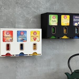 Op de afbeelding: Twee aan de muur gemonteerde thee-organizers, één witte en één zwarte, elk met drie verschillende theesoorten. De witte organizer toont thee met de labels "Hagebutte", "Blaubeere" en "Heiße Liebe". De zwarte organizer toont "Apfel", "Italianische Limone" en "Kamille".