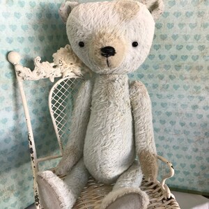 May include: A cream-coloured teddy bear with a vintage aesthetic, featuring a darker head and black button eyes. The bear sits on a white, ornate metal chair with a woven seat. The background is a light blue with a heart pattern.