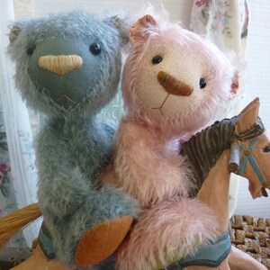 May include: Two plush teddy bears, one blue and one pink, ride a wooden rocking horse. The bears have stitched features and fluffy fur. The rocking horse is painted in muted tones with blue accents.