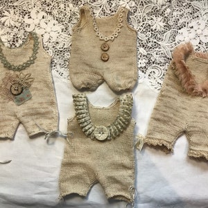 Puede incluir: Cuatro peleles de bebé de punto beige con escotes y adornos decorativos. Uno tiene un cuello de encaje, otro botones de madera y un tercero un cuello con volantes. El cuarto pelele tiene un adorno decorativo. Están dispuestos sobre una superficie blanca.