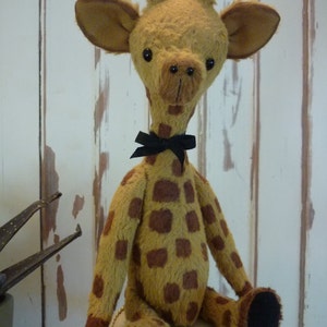 May include: A plush giraffe toy with a light brown body and dark brown spots. The giraffe has large ears, a long neck, and a black bow tie. It is sitting on a small, round, cream-colored seat.