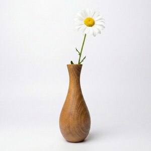 Puede incluir: Un jarr&oacute;n de madera con una sola margarita blanca. El jarr&oacute;n es de madera de color marr&oacute;n claro y tiene una forma suave y curvada. La margarita tiene un centro amarillo y p&eacute;talos blancos. El jarr&oacute;n mide unos 18 cm de altura.