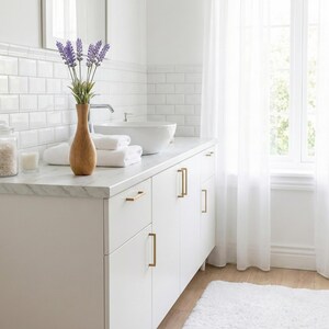 Puede incluir: Un ba&ntilde;o luminoso y limpio con un tocador blanco, encimera de m&aacute;rmol y herrajes dorados. Un jarr&oacute;n de madera contiene ramitas de lavanda. Toallas blancas, un tarro de cristal y una vela contribuyen al ambiente de spa. Cortinas blancas transparentes enmarcan una ventana.