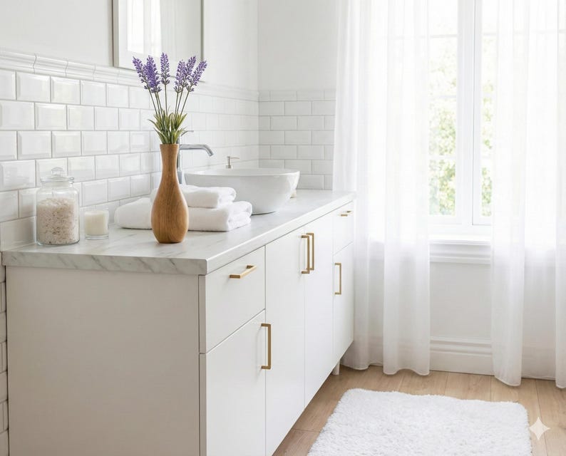 Puede incluir: Un ba&ntilde;o luminoso y limpio con un tocador blanco, encimera de m&aacute;rmol y herrajes dorados. Un jarr&oacute;n de madera contiene ramitas de lavanda. Toallas blancas, un tarro de cristal y una vela contribuyen al ambiente de spa. Cortinas blancas transparentes enmarcan una ventana.