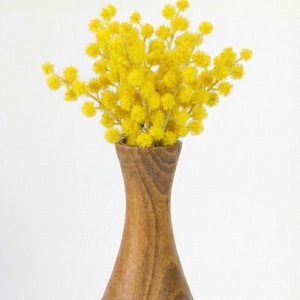 Puede incluir: Un jarr&oacute;n de madera con forma de l&aacute;grima contiene un vibrante ramo de flores amarillas. El jarr&oacute;n es de madera marr&oacute;n claro y tiene un acabado liso y pulido. Las flores son de un amarillo brillante y esponjoso, creando una exhibici&oacute;n alegre.