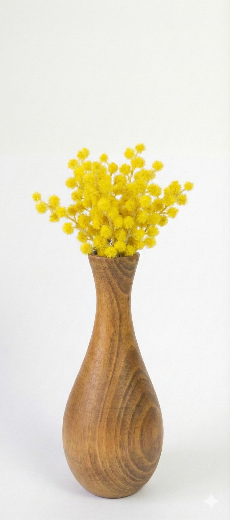 Puede incluir: Un jarr&oacute;n de madera con forma de l&aacute;grima contiene un vibrante ramo de flores amarillas. El jarr&oacute;n es de madera marr&oacute;n claro y tiene un acabado liso y pulido. Las flores son de un amarillo brillante y esponjoso, creando una exhibici&oacute;n alegre.