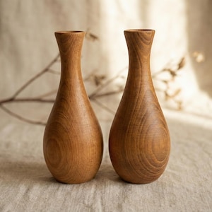 Puede incluir: Dos jarrones de madera con forma de l&aacute;grima y cuello estrecho. Los jarrones son de madera marr&oacute;n claro con vetas visibles. Se encuentran uno al lado del otro sobre una superficie de color neutro, con un fondo suave y natural.