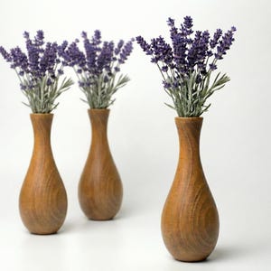 Puede incluir: Tres jarrones de madera con acabado marr&oacute;n claro, cada uno con un ramo de lavanda morada. Los jarrones tienen forma de l&aacute;grima con cuello estrecho. El fondo es blanco.