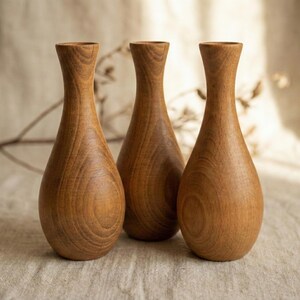 Puede incluir: Tres jarrones de madera con forma de l&aacute;grima y cuello estrecho. Los jarrones son de madera marr&oacute;n claro y tienen un patr&oacute;n de vetas naturales. Est&aacute;n dispuestos en fila sobre una superficie de color neutro.