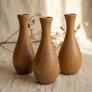 Puede incluir: Tres jarrones de madera con forma de l&aacute;grima y cuello estrecho. Los jarrones son de madera marr&oacute;n claro y tienen un acabado liso y pulido. Los jarrones est&aacute;n dispuestos en grupo sobre una superficie de color neutro.