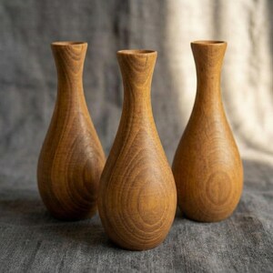 Puede incluir: Tres jarrones de madera con forma de l&aacute;grima y cuello estrecho. Los jarrones son de madera marr&oacute;n claro con vetas visibles. Los jarrones est&aacute;n dispuestos en formaci&oacute;n triangular sobre un fondo gris.