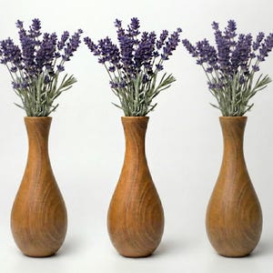 Op de afbeelding: Drie houten vazen, elk met een bosje lavendel. De vazen hebben een warme bruine kleur met een glad, gebogen ontwerp. De lavendel heeft paarse bloemen en groene stelen, wat een natuurlijk, decoratief arrangement creëert.