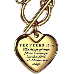 以下が含まれることがあります： 金色のネックレス、ハート型のペンダント付き。ペンダントには「PROVERBS 16:9」のテキストと引用句が書かれています。ネックレスはトグルクラスプと丸いリンクのチェーンが付いています。ハートペンダントの幅は約2.5cmです。