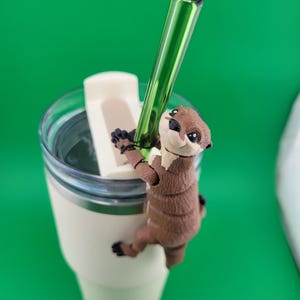 Może przedstawiać: Figurkę brązowej i beżowej wydry wspinającej się po boku białego kubka podróżnego z zieloną słomką szklaną wystającą z pokrywki.