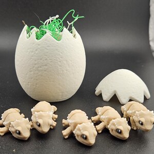 May include: Five small, tan, lizard-like figurines are hatching from white eggs. The eggs are cracked open and the figurines are in various poses.