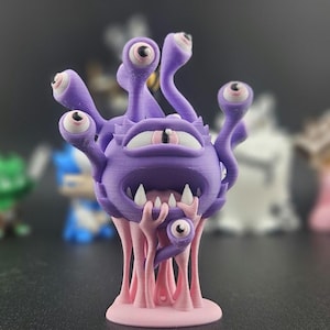 Puede incluir: Una figura de monstruo morado con múltiples ojos y tentáculos, de pie sobre una base rosa. La figura tiene una boca ancha y dientes afilados. Otras figuras están borrosas en el fondo.