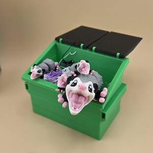May include: A green miniature dumpster with a black lid, filled with several gray and white opossum figurines. The opossums have pink noses and open mouths, and are surrounded by colorful string.