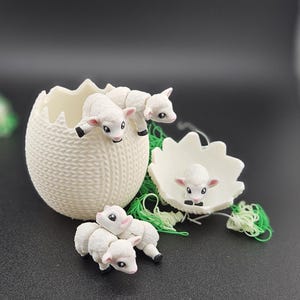 Könnte beinhalten: Ein weißer, eiförmiger Behälter aus Keramik mit einer Strickstruktur, der drei kleine weiße Plastiklämmer enthält. Ein Lamm befindet sich im Ei, zwei sind draußen, wobei eines aus dem Ei schaut.