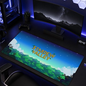 May include: A rectangular desk mat featuring a Stardew Valley graphic print. The design displays a vibrant green landscape with rolling hills, trees, and a blue sky with stars. The yellow text Stardew Valley is centred on the mat surface.