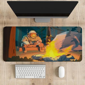 Pode incluir: Um mousepad retangular com um astronauta sentado perto de uma fogueira em um cenário de floresta. A ilustração tem uma paleta de cores quentes com chamas laranja e amarelas. Um teclado e um mouse de computador estão abaixo.