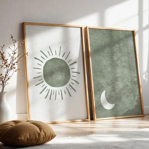 May include: Two framed art prints with a minimalist design. One features a green sunburst, the other a white crescent moon on a green background. Both are in light wood frames, leaning against a wall.