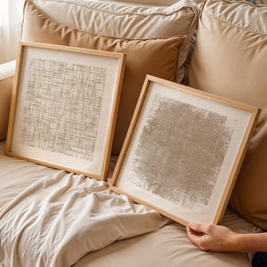 May include: Two framed abstract art prints with a neutral color palette. The artwork features a textured design in shades of beige and gray, set within light wood frames. The prints are displayed on a beige sofa with matching pillows.