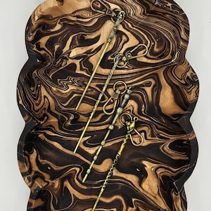 Puede incluir: Una bandeja de madera con un diseño veteado único en tonos marrón y negro. Varios llaveros de estilo antiguo con diseños de varita mágica están dispuestos en la bandeja, añadiendo un toque de fantasía.