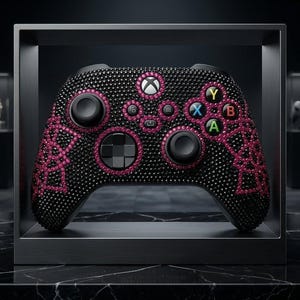 Puede incluir: Mando Xbox negro adornado con pedrería rosa y negra. El mando presenta detalles rosas alrededor de los botones y un diseño decorativo. El logotipo de Xbox es visible en el centro.