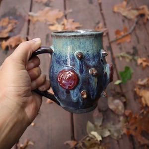 May include: A handmade ceramic mug with a unique design. The mug features a dark blue and teal glaze with raised, textured elements in brown and red. The mug is held by a hand, showcasing its handle and overall shape.