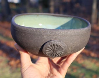 May include: A handmade ceramic bowl with a dark gray exterior and a light green interior. The bowl features a carved floral design on the side. The bowl is held in a hand against a blurred outdoor background.