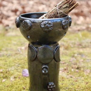 May include: A dark green ceramic incense burner with a round bowl and a tall, cylindrical base. The bowl has decorative floral accents, and the base features crescent moon and floral designs. A bundle of dried herbs rests inside.