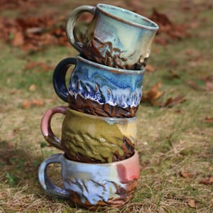May include: A stack of four handmade ceramic mugs with textured, earthy designs. The mugs feature a variety of colors, including blue, green, and brown, and are stacked on top of each other. The mugs are on a bed of grass and fallen leaves.