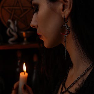 Può includere: Una persona indossa un orecchino a forma di mezzaluna con una catena pendente e una collana abbinata. Sta tenendo una candela accesa. Lo sfondo include un pentagramma e altri simboli occulti, creando un'estetica gotica.