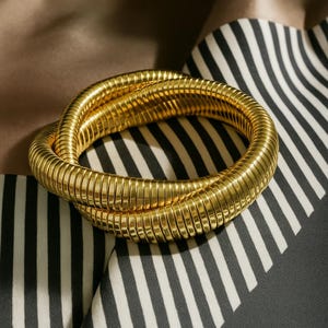 Peut inclure: Un bracelet doré enroulé, avec une texture côtelée. Le bracelet est entrelacé et repose sur un fond de tissu à rayures noires, blanches et beige.