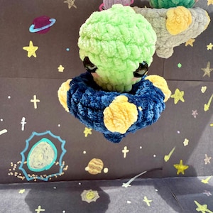 May include: A green crocheted alien with black eyes and a blue crocheted flying saucer with yellow accents. The alien is hanging from the ceiling against a black background with white stars and planets drawn on it.