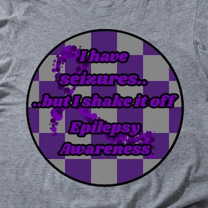 May include: A gray t-shirt with a circular graphic promoting epilepsy awareness. The graphic features a purple and gray checkered pattern with the text "I have seizures...but I shake it off" and "Epilepsy Awareness".