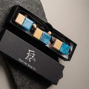 May include: A black box with the text "Dont Die Soap Co" and a logo containing a plague doctor. Inside the box are three wooden and blue resin cube-shaped pendants on black cords. The box is on a dark gray stone.