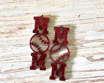 May include: Red glitter baseball earrings with the word "MOM" in a bold font. The "O" is replaced with a baseball design. The earrings are hanging against a light wood grain background.