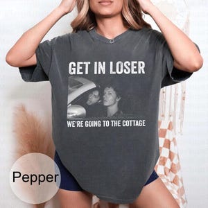 May include: Pepper-colored t-shirt with the text "GET IN LOSER" above a black and white image of two people in a car. Below the image, the text reads "WE'RE GOING TO THE COTTAGE".