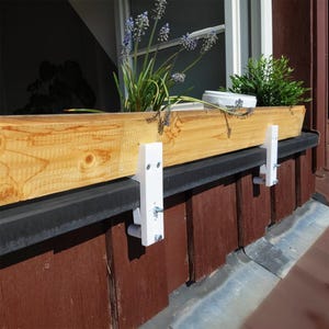 Könnte beinhalten: Ein hölzerner Blumenkasten mit Pflanzen, der von weißen Halterungen getragen wird. Die Box besteht aus hellem Holz und ist mit grünen Pflanzen und lila Blumen gefüllt. Die Halterungen sind am Fensterrahmen befestigt.