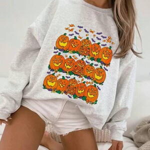 Pode incluir: Uma camisola de gola redonda cinzento-claro com uma impressão gráfica de várias abóboras cor de laranja com rostos esculpidos, intercaladas com pequenos morcegos roxos e formas de milho doce cor de laranja num padrão retangular.