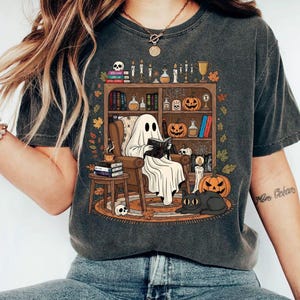 Könnte beinhalten: Dunkelgraues T-Shirt mit einer Halloween-Illustration. Ein Geist liest in einem Sessel vor einem Bücherregal mit Kürbissen, Kerzen und einer schwarzen Katze. Das Design umfasst einen Totenkopf, Bücher und Herbstblätter.
