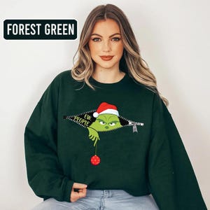 May include: Forest green sweatshirt featuring a graphic of a green character wearing a Santa hat, peeking out of a zipper. The text "EW. PEOPLE" is visible. The character is holding a red ornament. The sweatshirt is a solid forest green color.