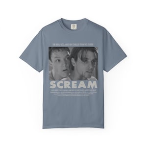 Pu&ograve; includere: Una maglietta blu-grigia con un'immagine in bianco e nero del film "Scream". L'immagine mostra due persone e il titolo "SCREAM" in bianco. Il testo "THE HIGHLY ACCLAIMED NEW THRILLER FROM WES CRAVEN" &egrave; sopra l'immagine.
